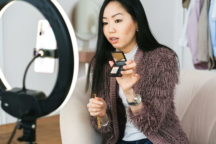 Young woman applying makeup on camera with ring light, illustrating trends leading to overconsumption in beauty content creation.