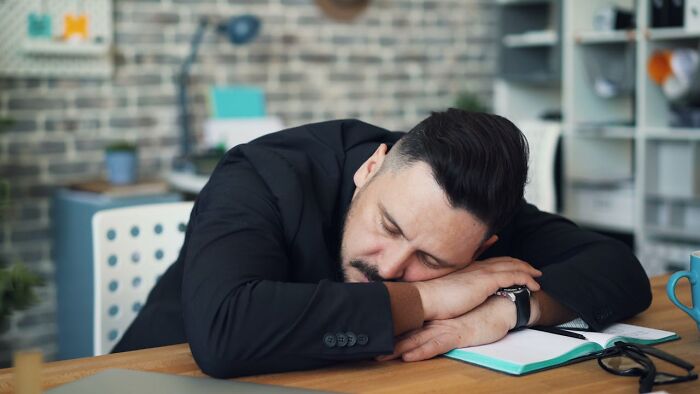 Man in black suit taking revenge on bosses concept, resting head on crossed arms over notebook at office desk.