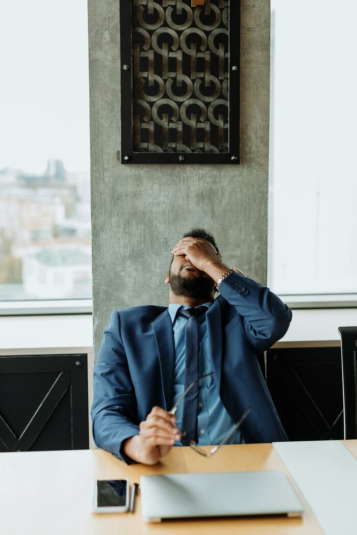 Stressed man in blue suit at office desk, holding glasses and covering face, reflecting workplace revenge and frustration.