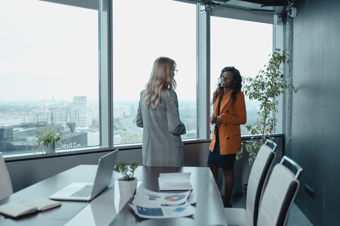 Two professional women discussing workplace issues by a window in an office, reflecting on revenge on their bosses.