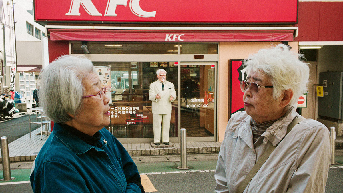 Two elderly women in conversation outside a KFC in Japan capturing daily life moments by Shin Noguchi.