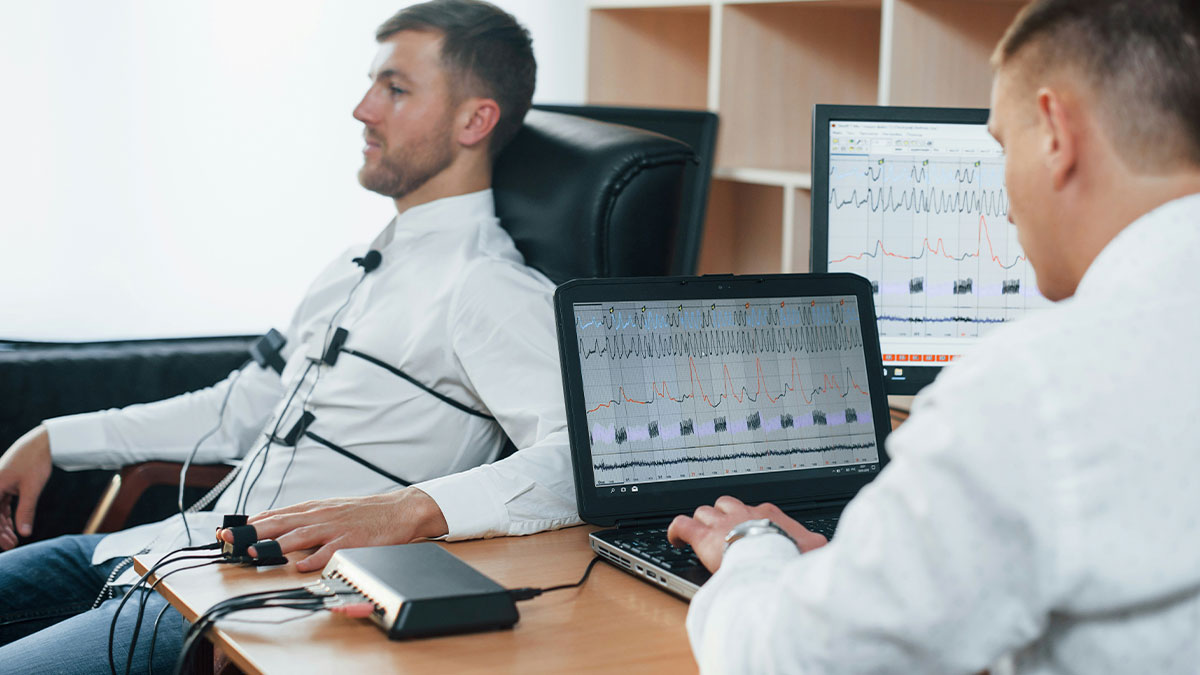 Man undergoing polygraph test while technician monitors bizarre beliefs and myths data on laptop screens.