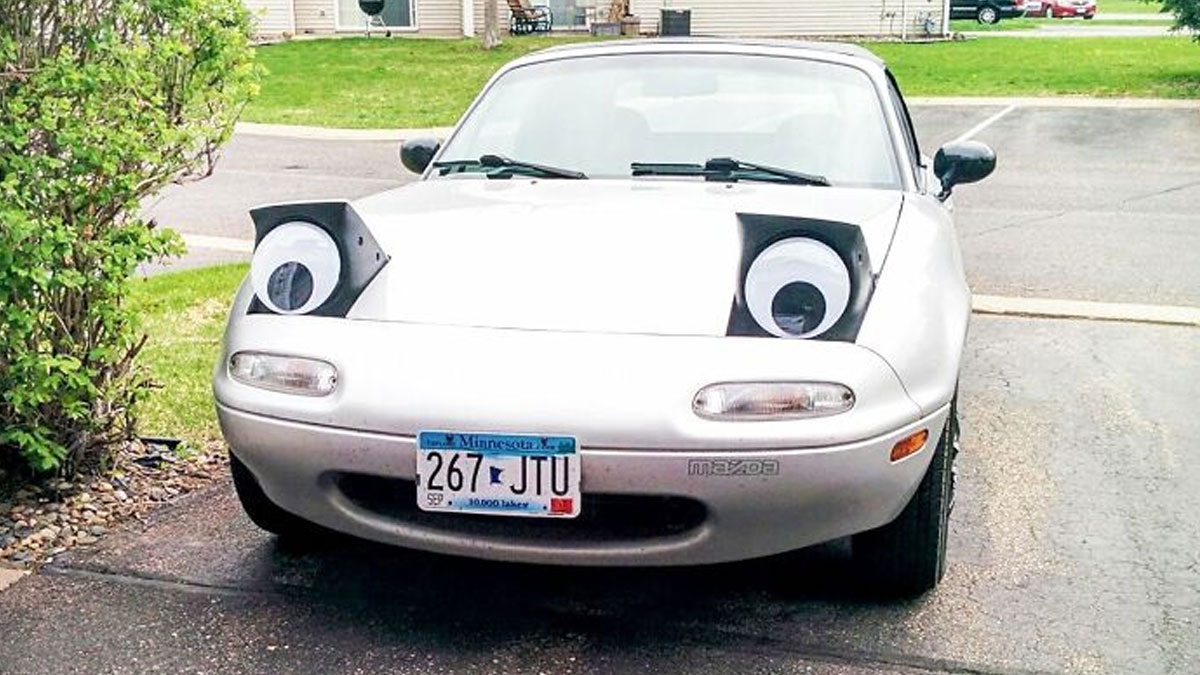 White Mazda car with quirky oversized googly eyes attached to headlights parked in a driveway outdoors