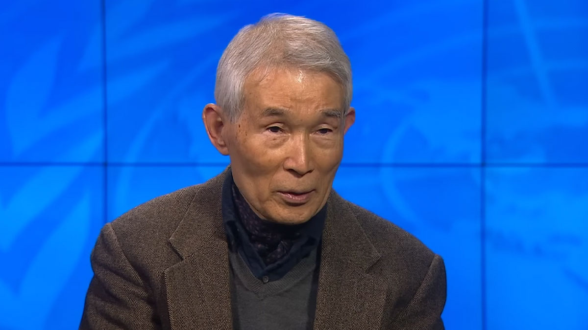 Elderly man in a brown jacket discussing crazy but real historical events in a TV studio with a blue background.
