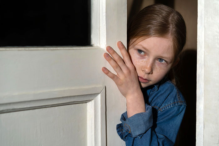 Young girl in a denim shirt looking anxiously around a doorframe, reflecting fears about autistic brother's safety concerns.