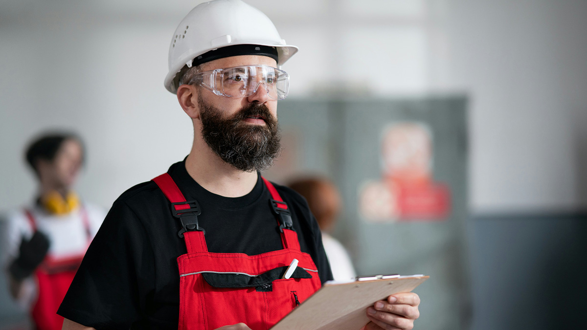 Male employee in a hard hat and safety glasses holding a clipboard, possibly reacting to wage reduction at work.