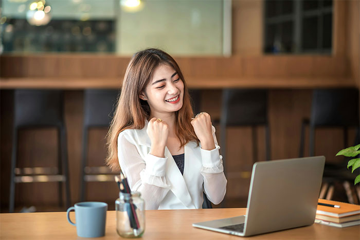 Young woman with PhD celebrating success at laptop in modern indoor workspace with coffee cup and notebooks on table Young woman with PhD celebrating success at laptop in modern indoor workspace with coffee cup and notebooks on table