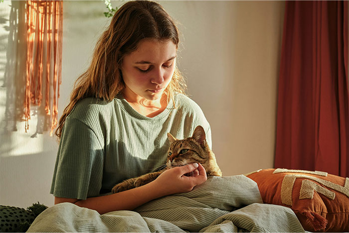 Woman sitting on bed holding and looking at her cat, appearing contemplative in a softly lit room Woman sitting on bed holding and looking at her cat, appearing contemplative in a softly lit room