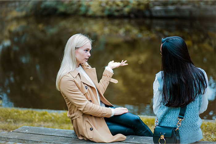 Two women sitting outdoors near water, one angrily explaining while the other listens, highlighting relationship conflict. Two women sitting outdoors near water, one angrily explaining while the other listens, highlighting relationship conflict.