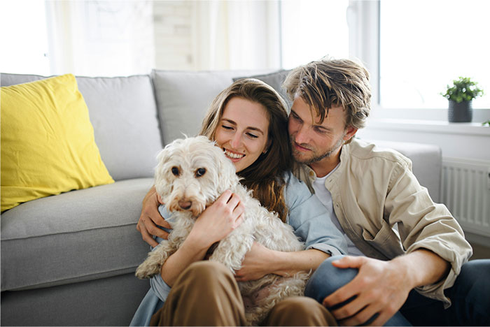 Couple sitting on floor with dog, woman smiling while man looks at her, representing woman furious over cat renaming. Couple sitting on floor with dog, woman smiling while man looks at her, representing woman furious over cat renaming.