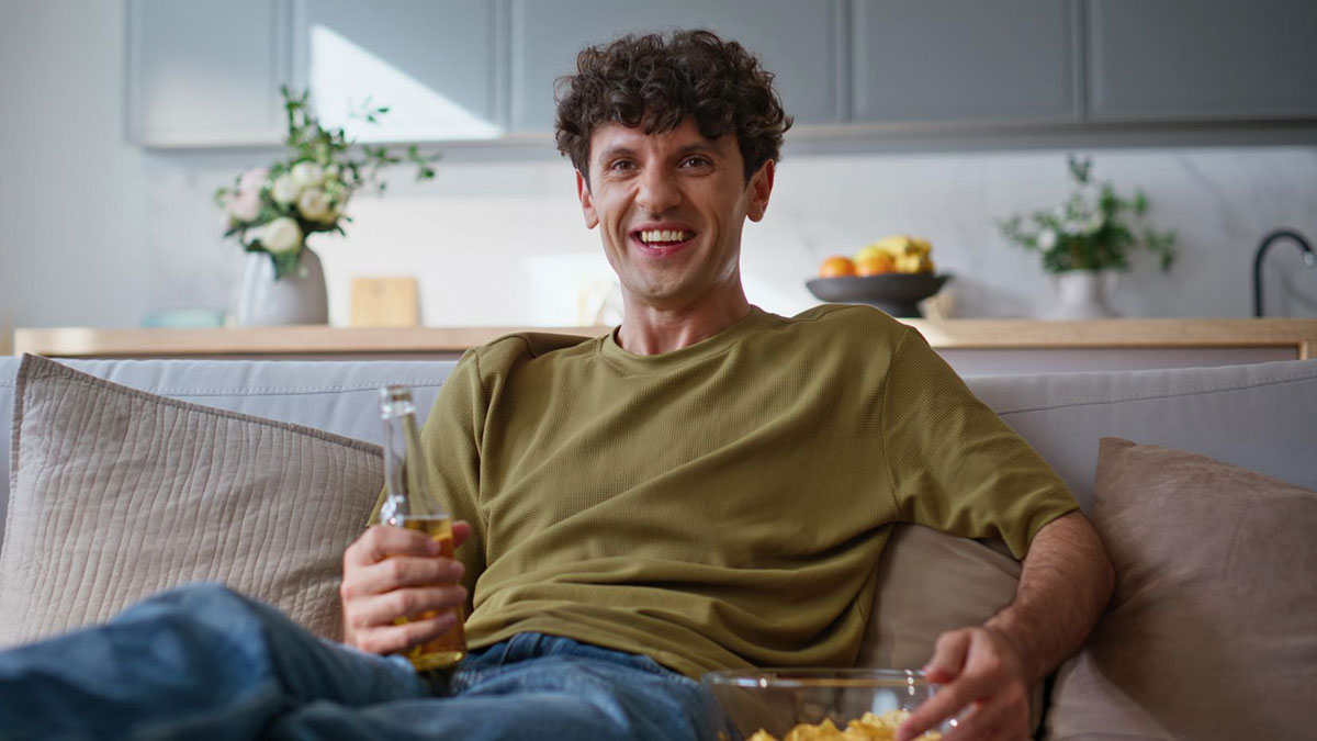 Young man relaxing on couch with drink and snacks, reacting to woman who doesn't believe in dinosaurs controversy.