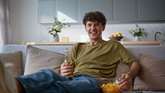 Young man relaxing on couch with beer and chips while watching Jurassic Park, a woman nearby expressing disbelief in dinosaurs. Young man relaxing on couch with beer and chips while watching Jurassic Park, a woman nearby expressing disbelief in dinosaurs.
