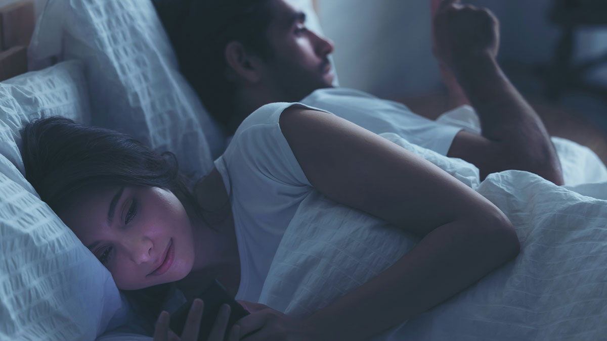 Woman in bed smiling at phone while man lies awake, illustrating outrageous student names shared by a teacher