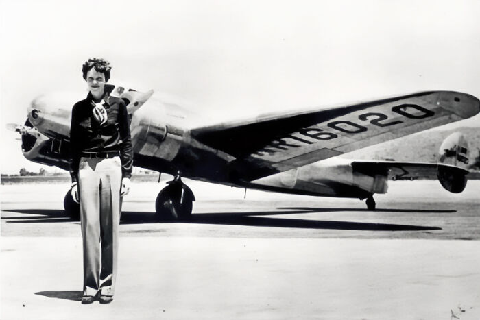 Amelia Earhart standing beside her Lockheed Electra plane in a vintage black and white photograph at an airfield. Amelia Earhart standing beside her Lockheed Electra plane in a vintage black and white photograph at an airfield.