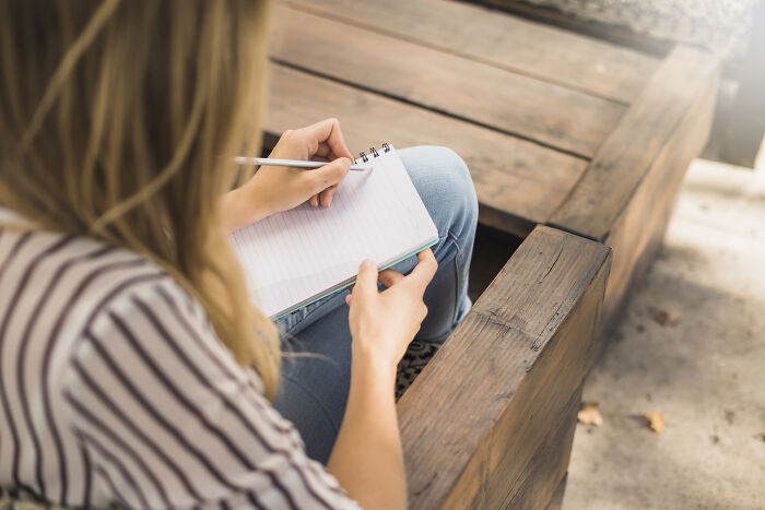 Woman sitting outdoors on wooden bench, writing in notebook, reflecting on sentences that changed people's lives.