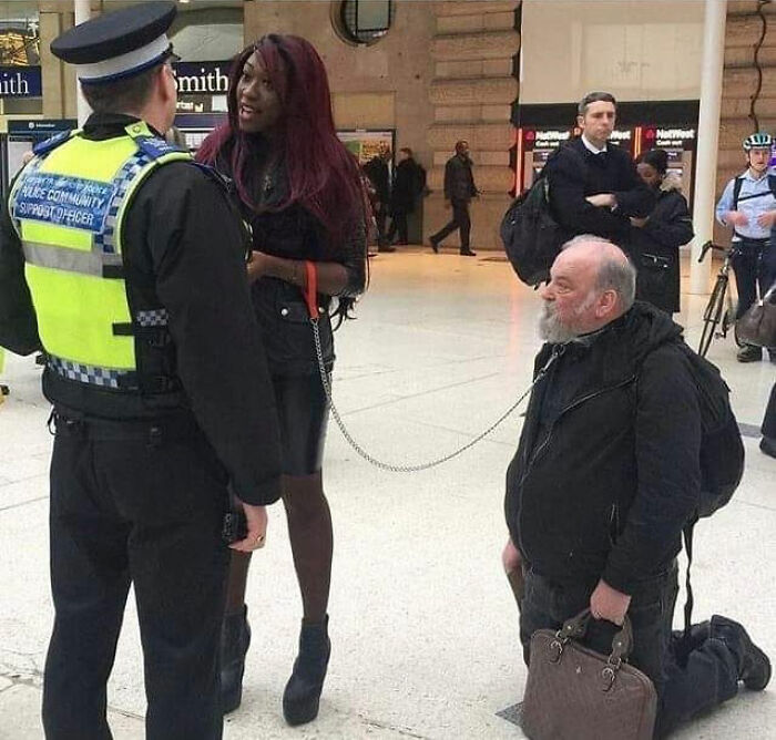 Man on leash held by woman talking to police officer in crowded indoor public space, a cursed image scenario.