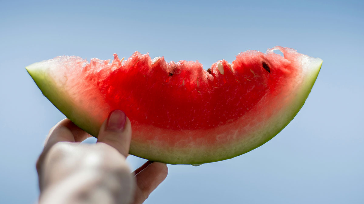 Hand holding a partially eaten slice of watermelon against a clear blue sky showing side effects of eating watermelon.