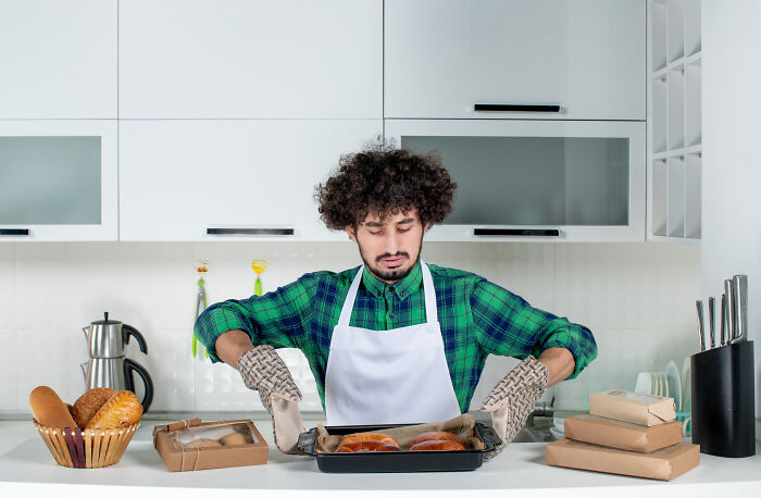 Man wearing oven mitts and apron baking bread in kitchen, showing signs grew up sheltered in a home environment