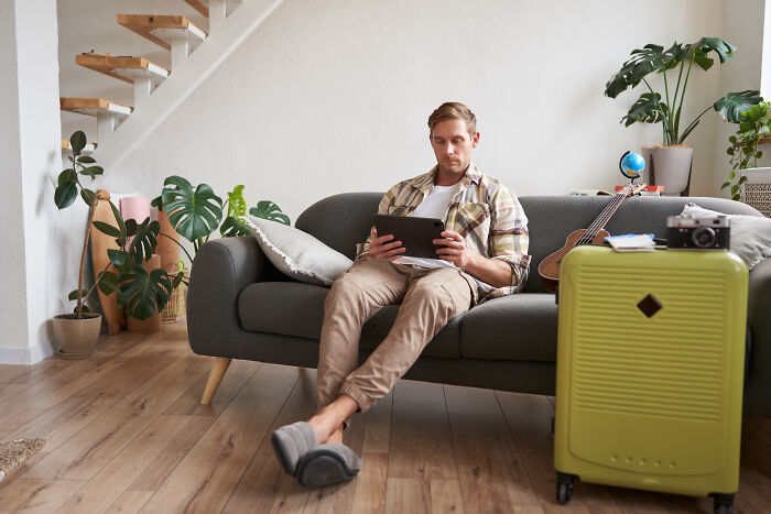 Young man sitting on sofa at home using a tablet, surrounded by plants and a green suitcase, signs grew up sheltered.