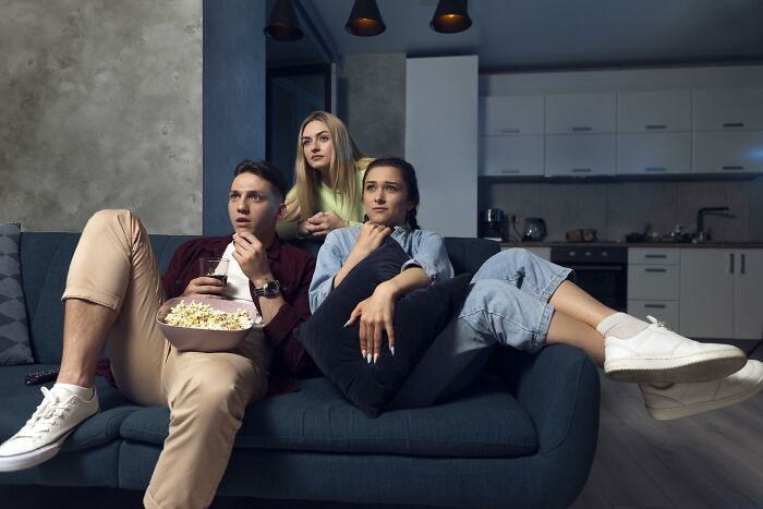 Three young adults watching TV on a couch at night, showing signs grew up sheltered with cautious expressions.