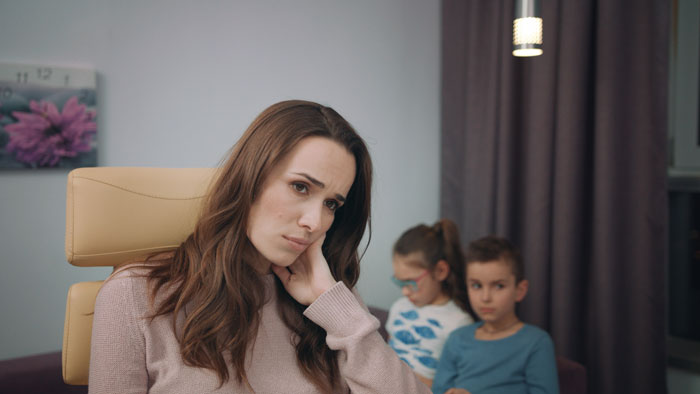 Worried woman sitting in chair while two children in background, illustrating family conflict and late wife impact. Worried woman sitting in chair while two children in background, illustrating family conflict and late wife impact.
