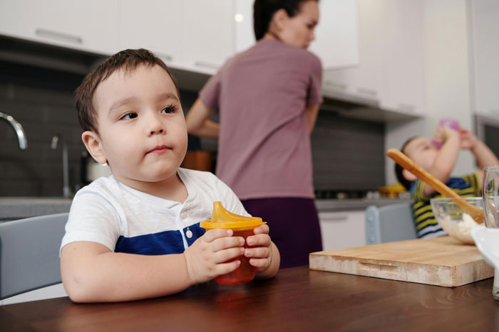 Young child with sippy cup sitting at the table while mother busily attends to kitchen tasks in background. Young child with sippy cup sitting at the table while mother busily attends to kitchen tasks in background.