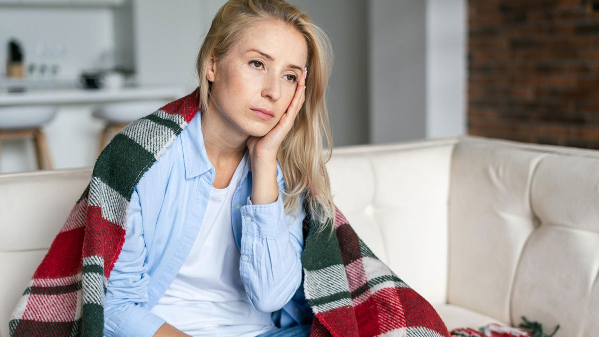 Woman wrapped in blanket sitting on couch, looking sad and contemplative, reflecting on infertility and relationship struggles.