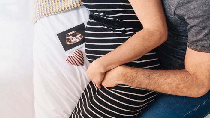 Pregnant woman in striped dress embraced by partner with ultrasound photo on bed, symbolizing baby and conception struggles. Pregnant woman in striped dress embraced by partner with ultrasound photo on bed, symbolizing baby and conception struggles.