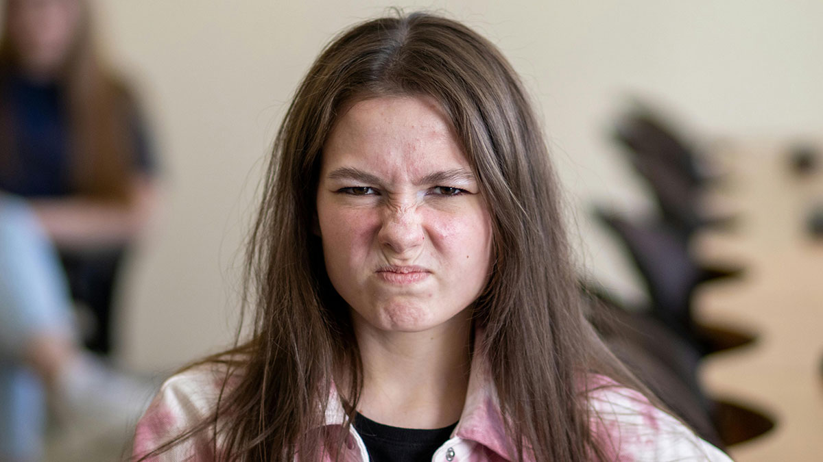 Angry teenage girl with long hair in a classroom setting expressing frustration with sibling behavior.