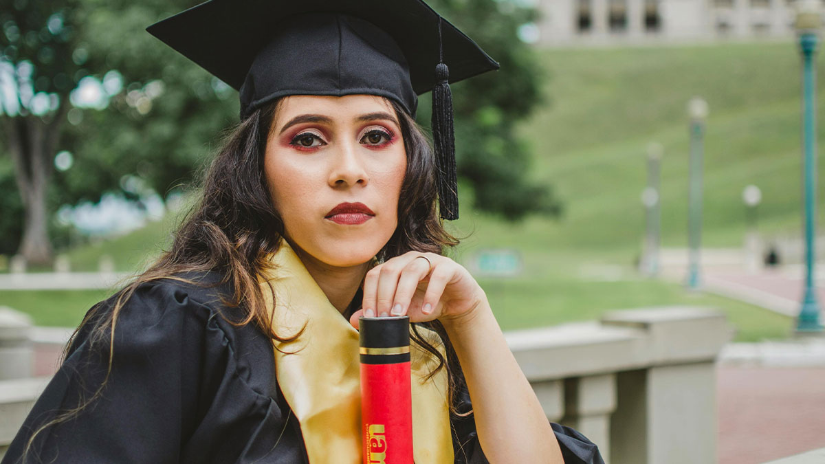 Woman in graduation gown and cap holding diploma, looking upset on her graduation day outside campus area.