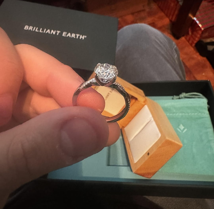 Hand holding a small girlfriend proposal ring with a diamond, next to an open wooden ring box and Brilliant Earth packaging.