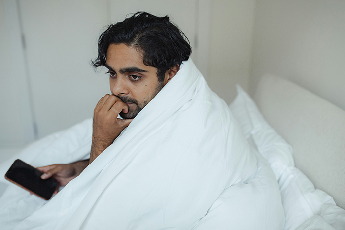 Man wrapped in a white blanket sitting on bed looking pensive while holding a phone, reflecting on small propose ring choice