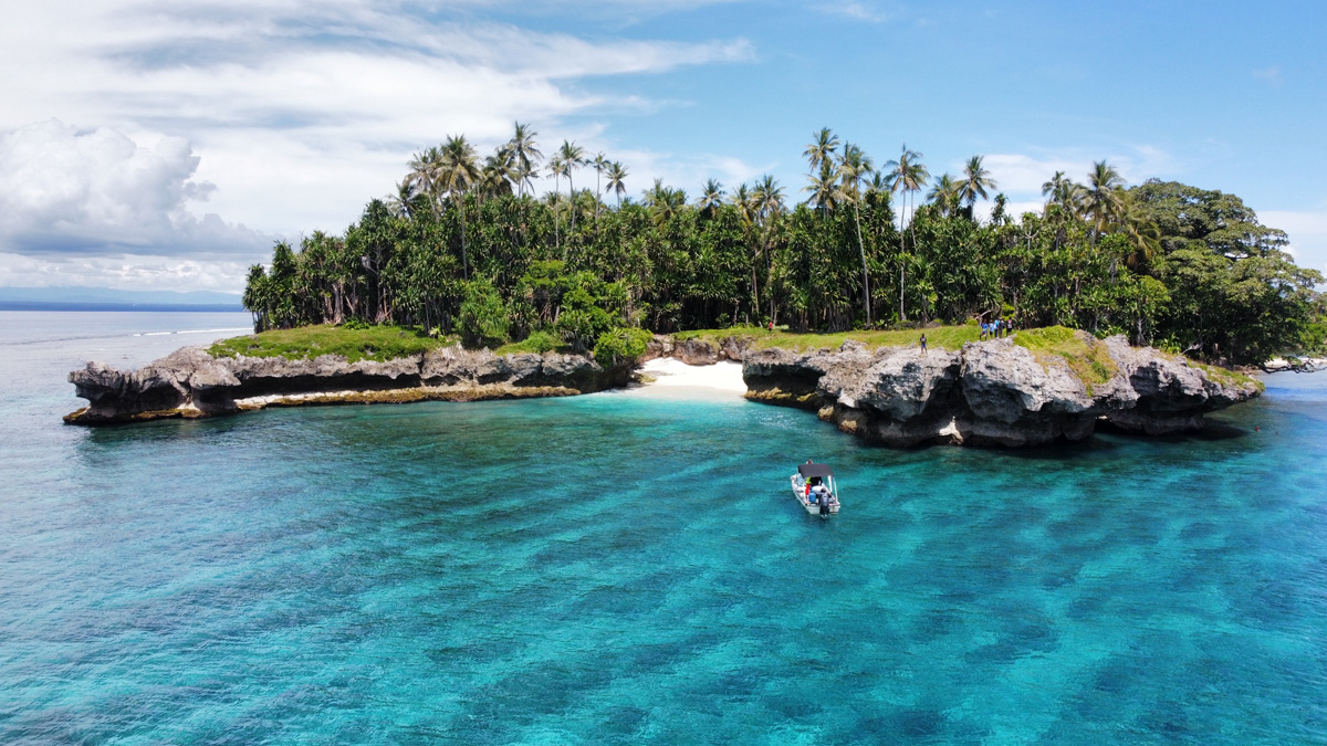 A small boat near a lush tropical island with a rocky shore, related to a cannibal tribe skull cave trek in Papua New Guinea.
