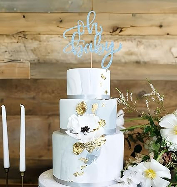 Elegant three-tier custom baby shower cake with gold accents and floral decorations on a rustic wooden background.