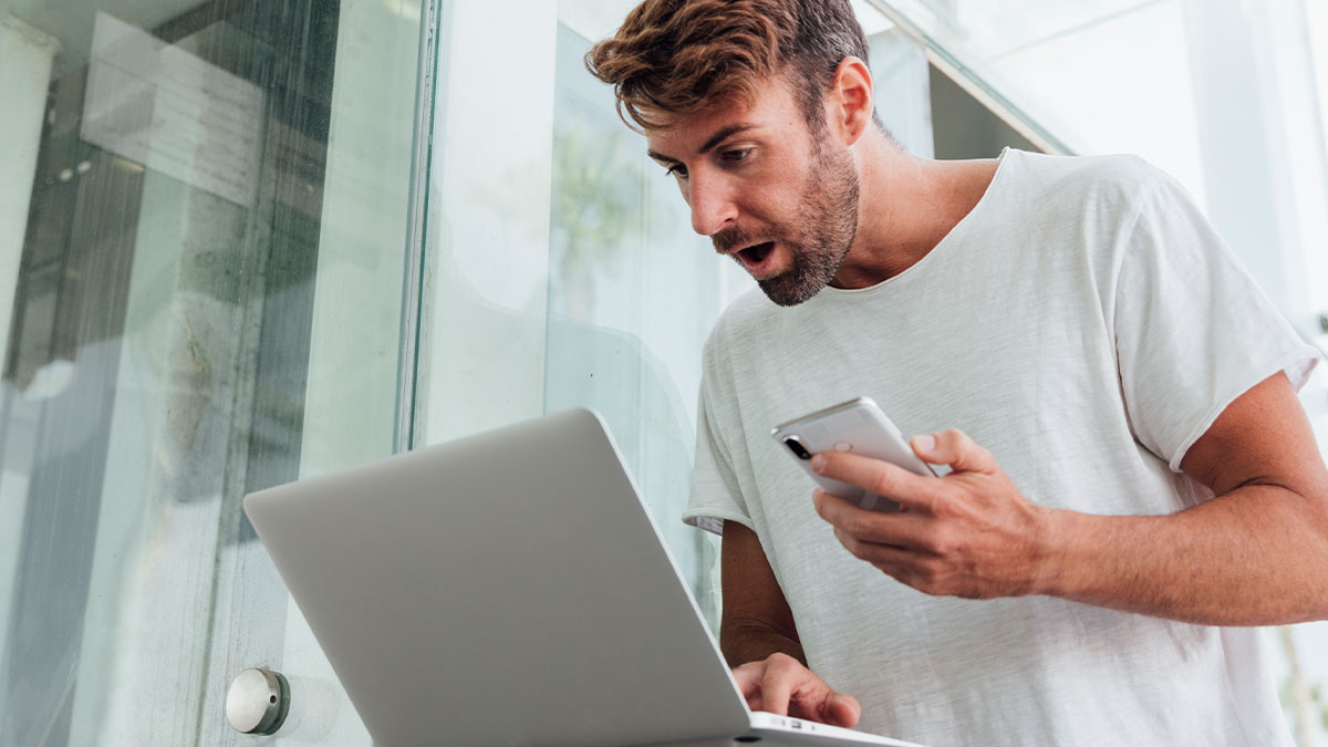 Man looking shocked at laptop screen while holding phone, reacting to sending something to wrong person stories