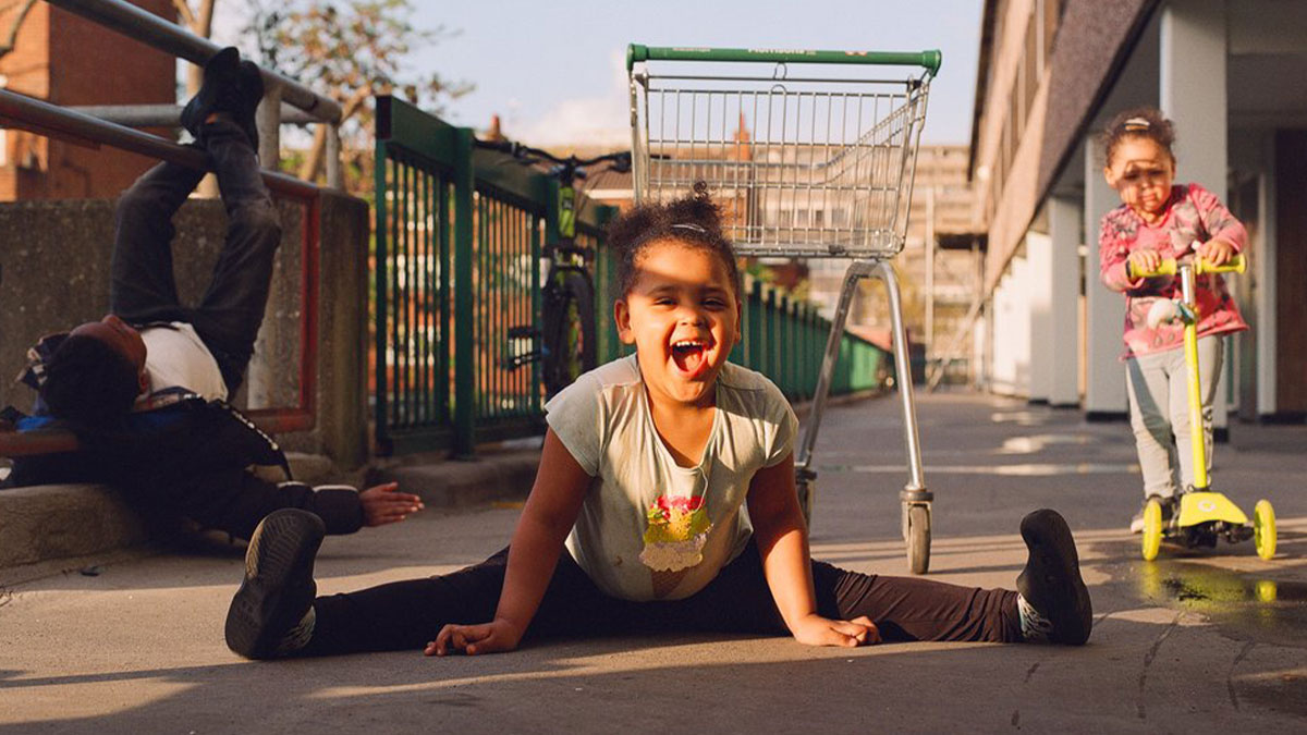 Children playing on a London street with a shopping trolley, captured in an honest photo by Nico Froehlich.