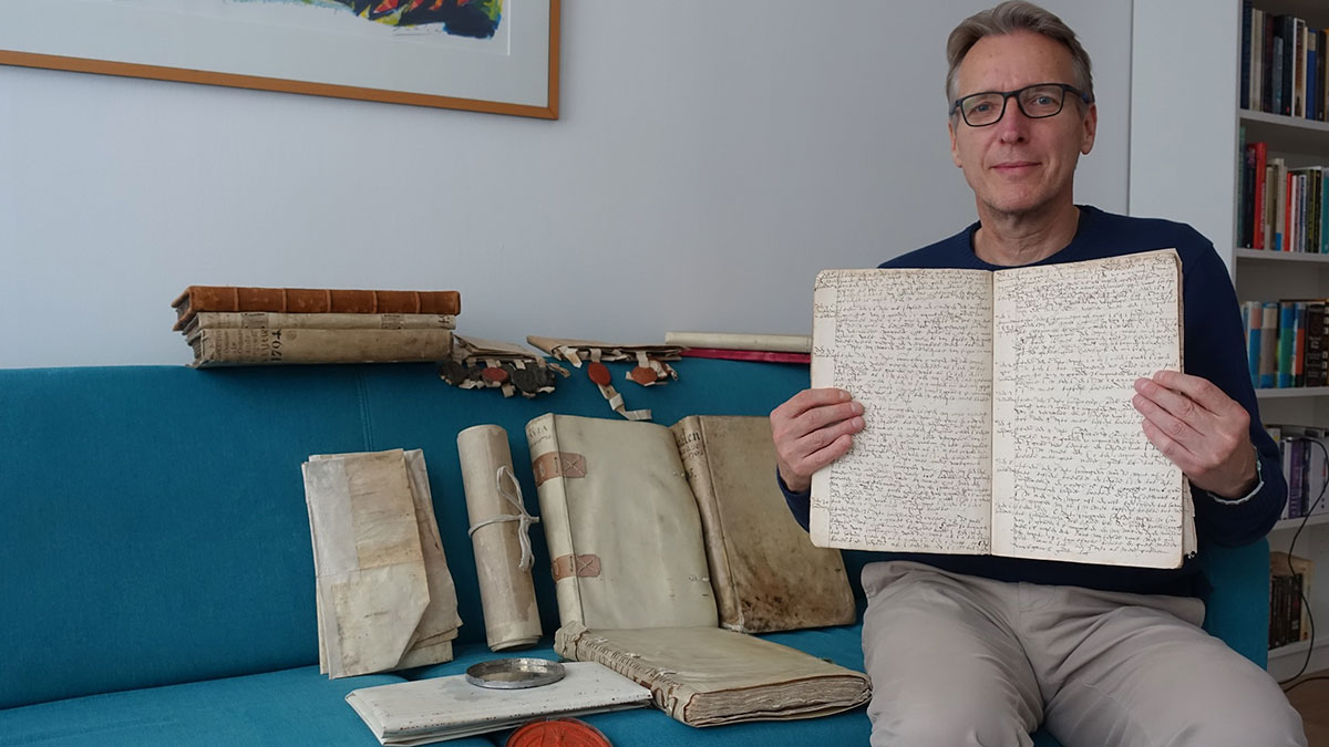 Man wearing glasses holding historic documents on sofa with several old books and manuscripts displayed around him