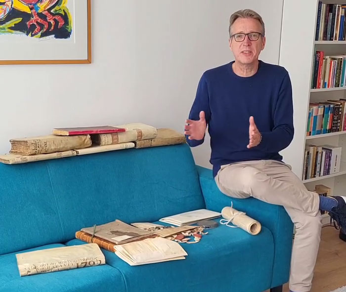 Man in glasses sitting next to stolen historic documents spread on a blue couch, discussing the documents' resurfacing. Man in glasses sitting next to stolen historic documents spread on a blue couch, discussing the documents' resurfacing.