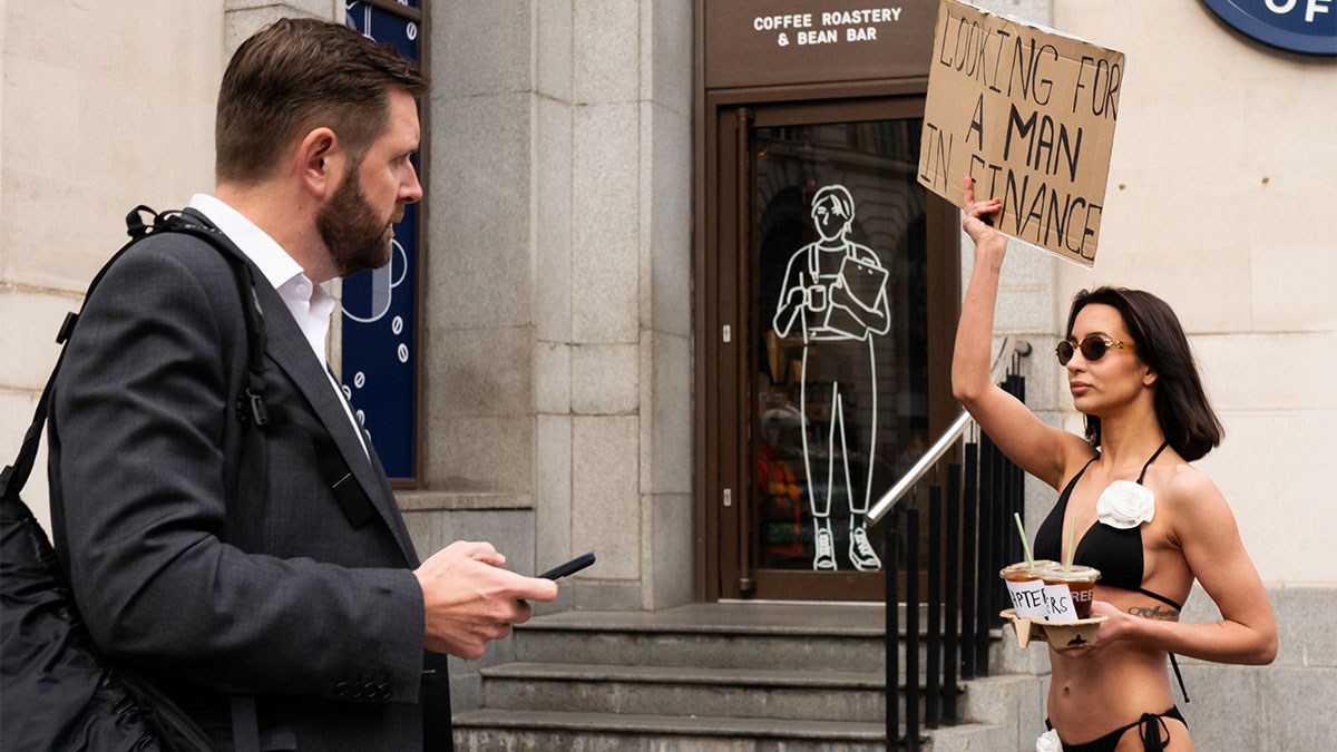 Woman in bikini holding sign and coffee cups while man in suit looks on, a coincidental moment captured by photographer in public.