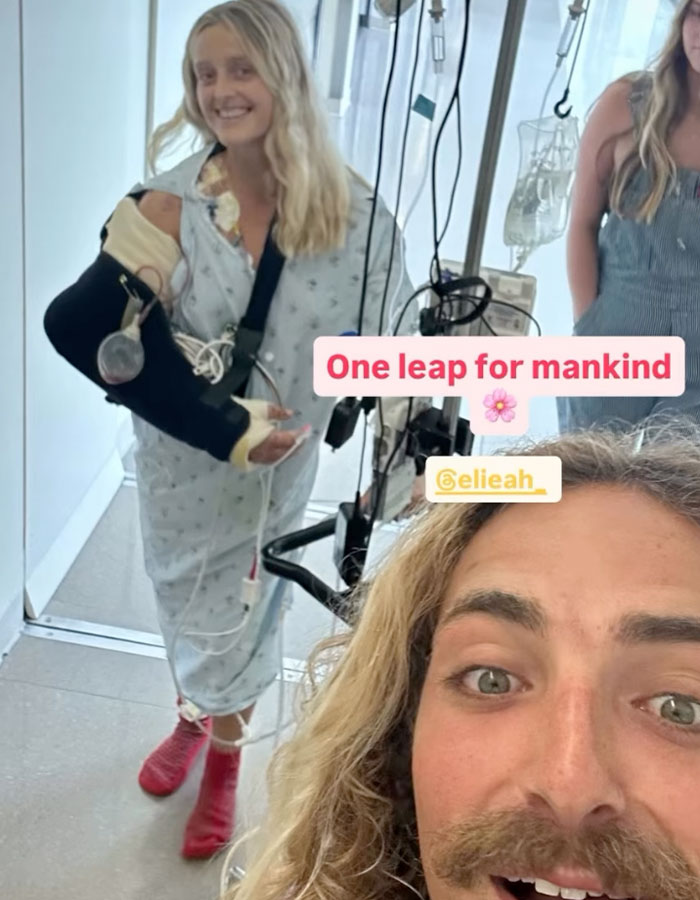 Young surf instructor in hospital gown with arm in a cast and sling, smiling during recovery after train accident. Young surf instructor in hospital gown with arm in a cast and sling, smiling during recovery after train accident.