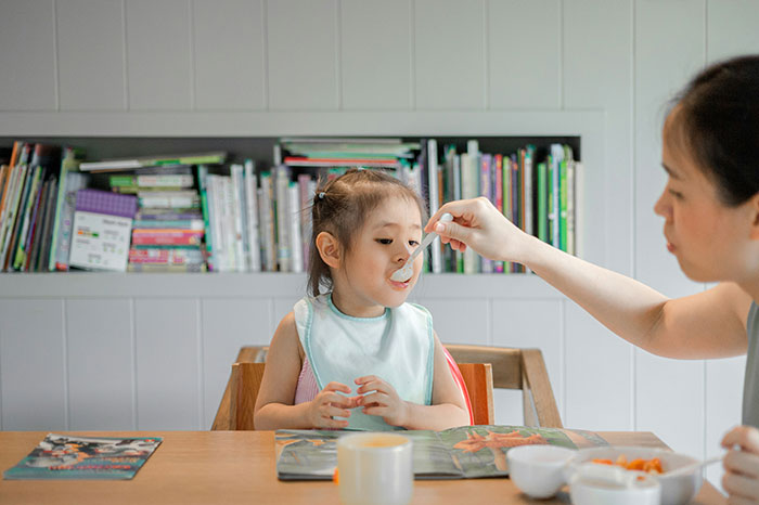 Young adult woman feeding half-sibling at the table, illustrating responsibility of caring for younger family members.