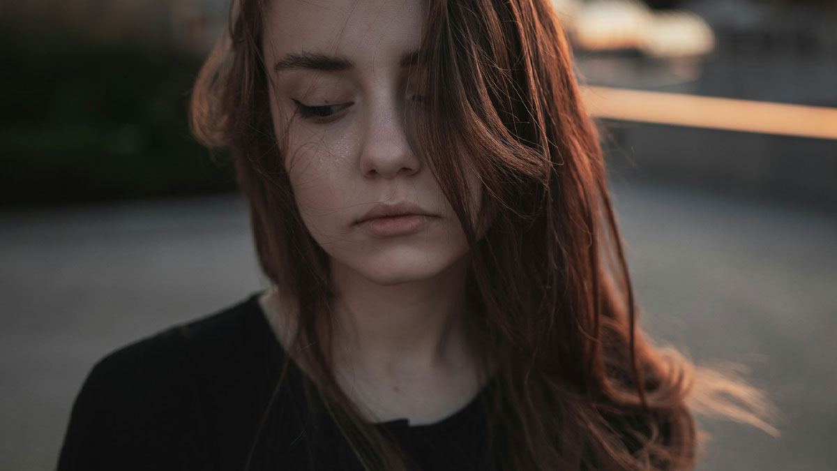 Sad adopted daughter looking down outdoors, reflecting on parents playing favorites after graduation and moving out.