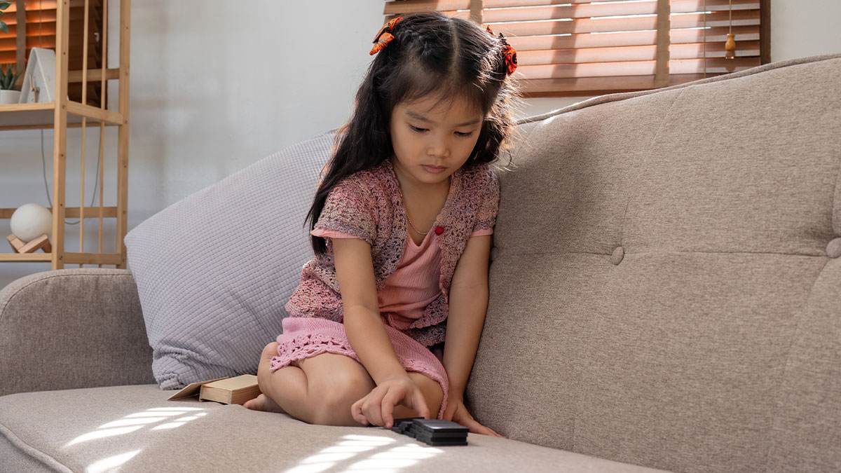 Young girl left alone on couch playing with remote control, highlighting issues of babysitting emergency neglect.