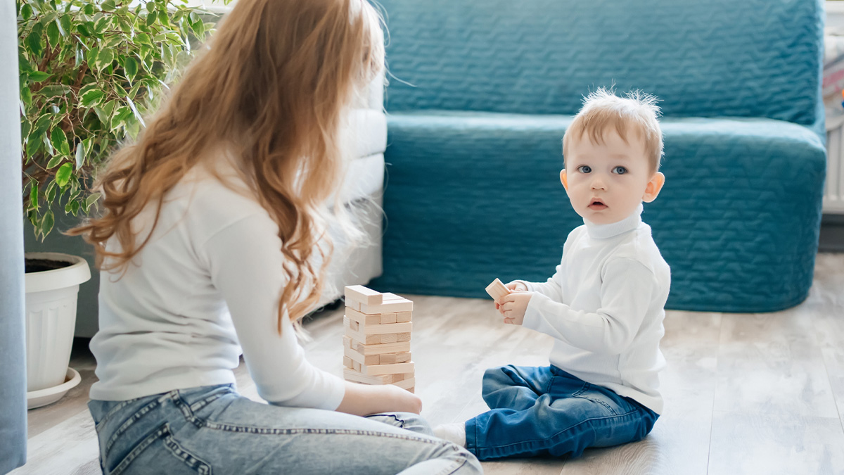 Woman babysitting toddler playing with wooden blocks indoors near couch and plant in bright living room