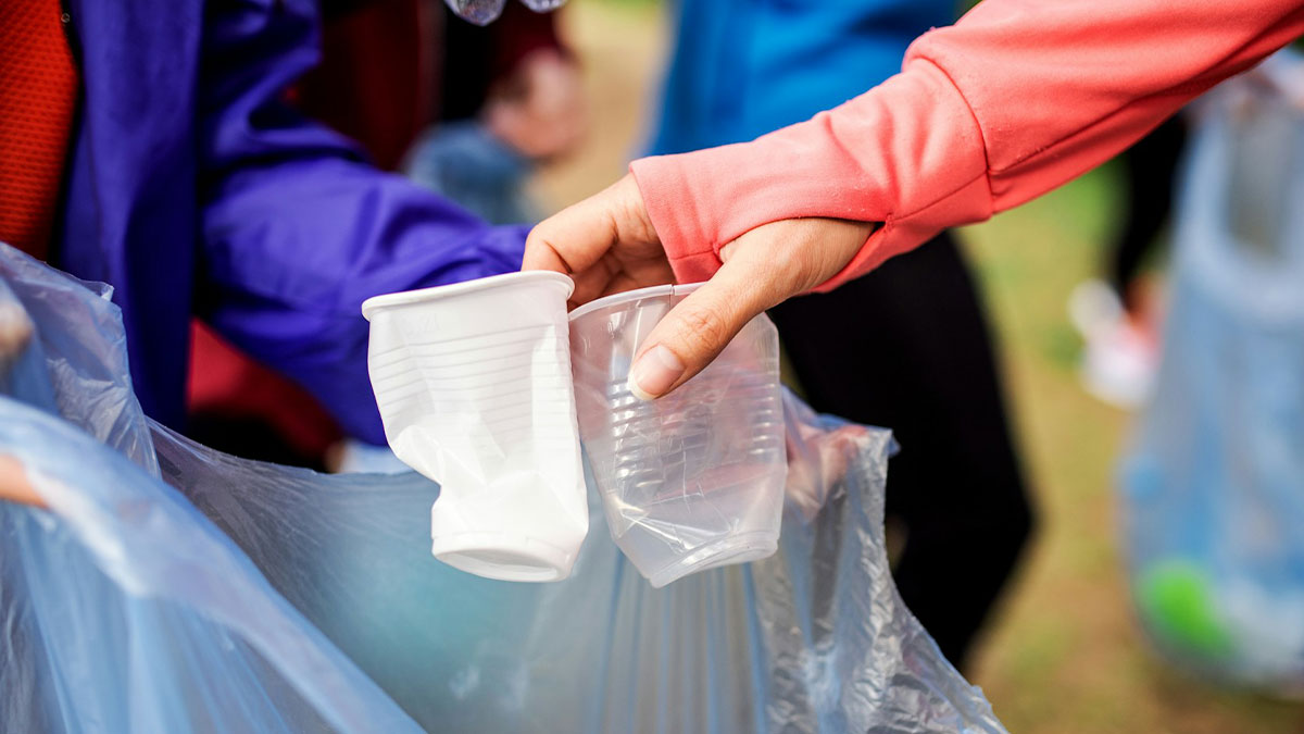 Person wearing a red sleeve holding crushed plastic cups over a trash bag, illustrating waste and environmental collapse issues.
