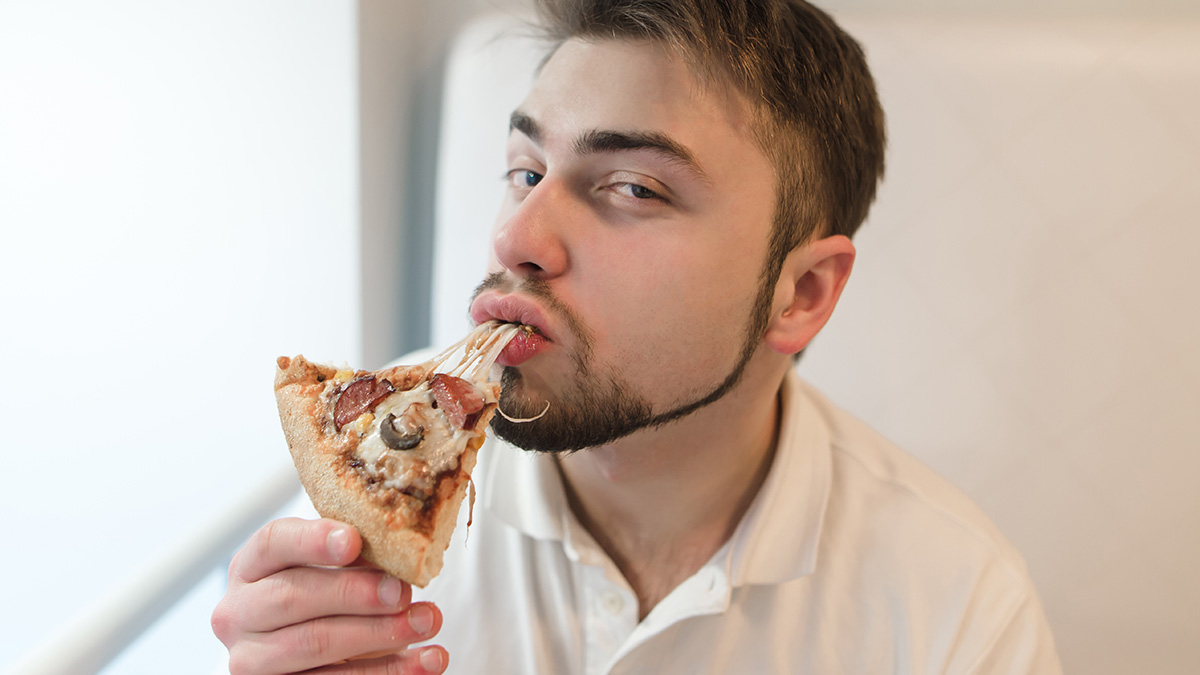 Young man eating pizza slice with melted cheese, illustrating relatable facts people are tired of explaining to others.