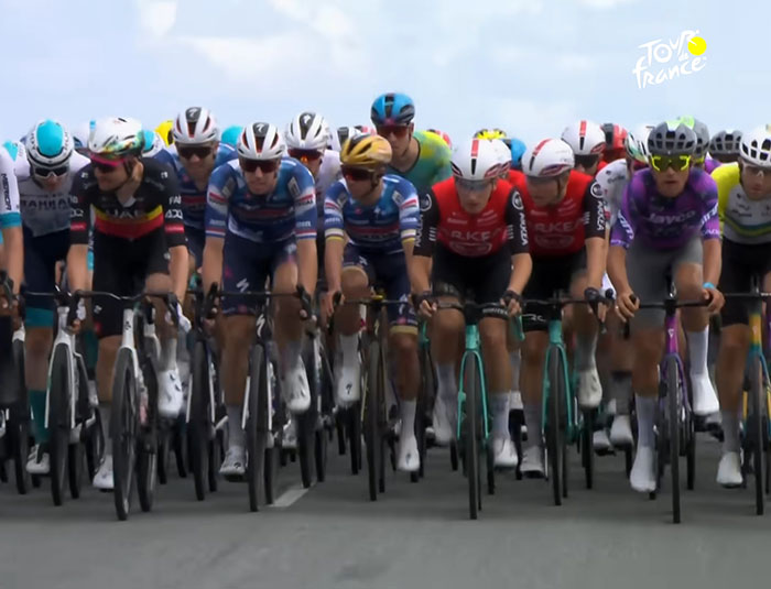 Tour de France cyclists ride closely in a group during a race, with fans controversially standing on graves nearby. Tour de France cyclists ride closely in a group during a race, with fans controversially standing on graves nearby.