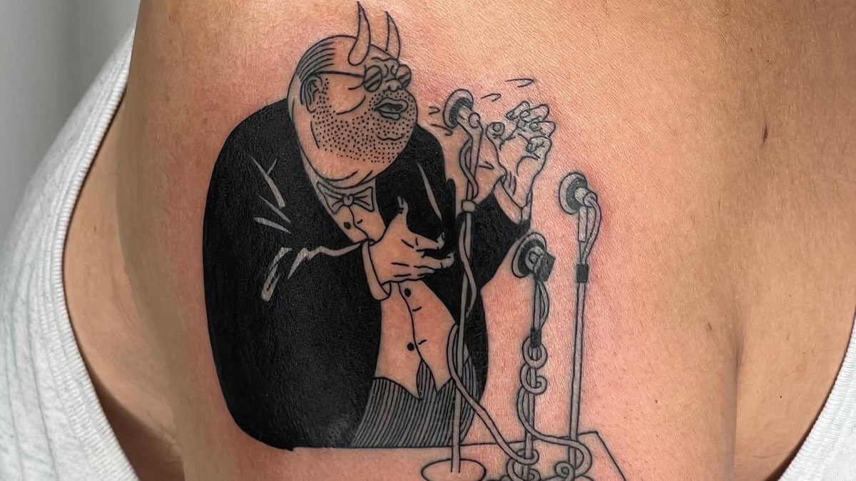 Tattoo of a man in a tuxedo with devil horns speaking into multiple vintage microphones, detailed black and gray ink work.