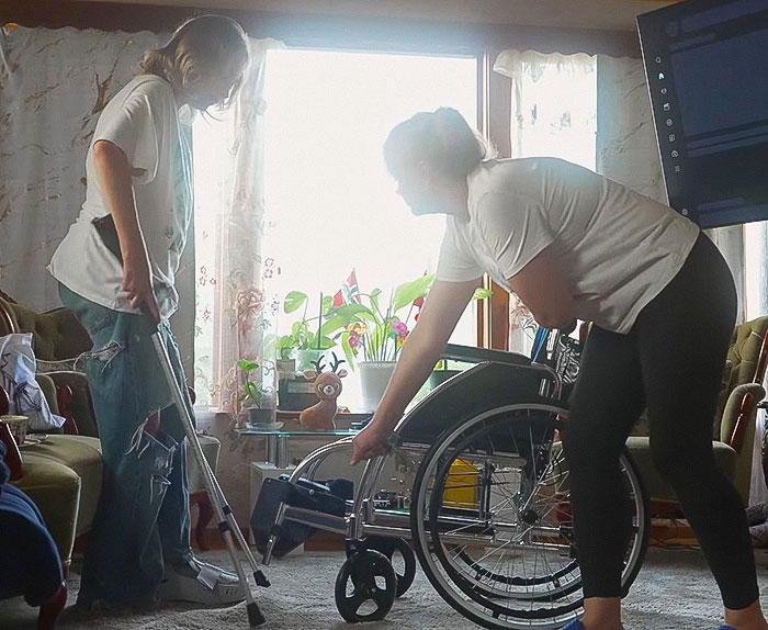 Person with broken limbs using crutches assisted by another person near a wheelchair in a cozy living room setting. Person with broken limbs using crutches assisted by another person near a wheelchair in a cozy living room setting.