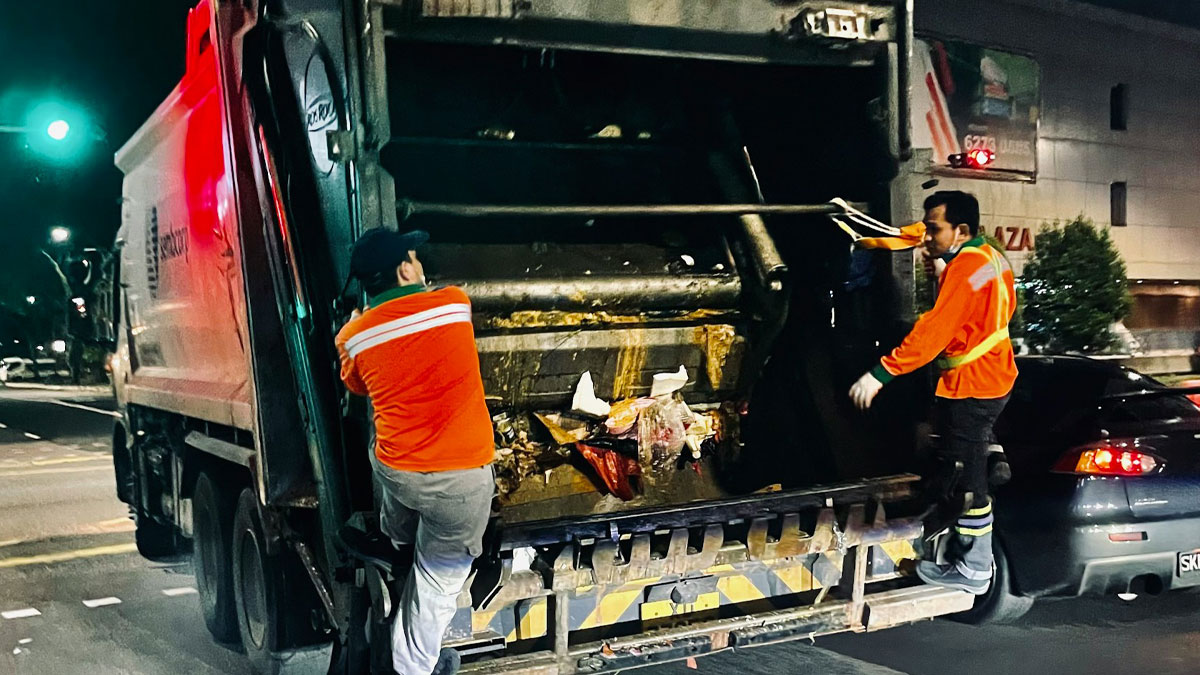 Two sanitation workers in reflective orange jackets collecting trash from a garbage truck at night on city street.